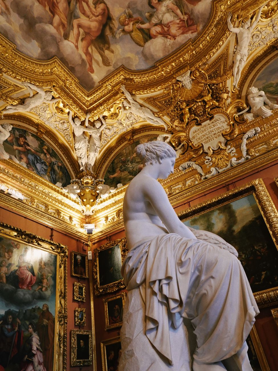 A marble statue of a woman seated in an ornate room, featuring elaborate gold-detailed ceiling artwork and framed paintings on the walls.