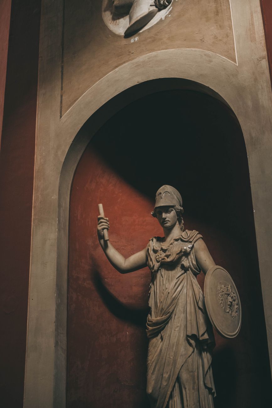 A marble statue of a warrior figure holding a scroll in one hand and a shield in the other, set against a reddish wall in an arched niche.